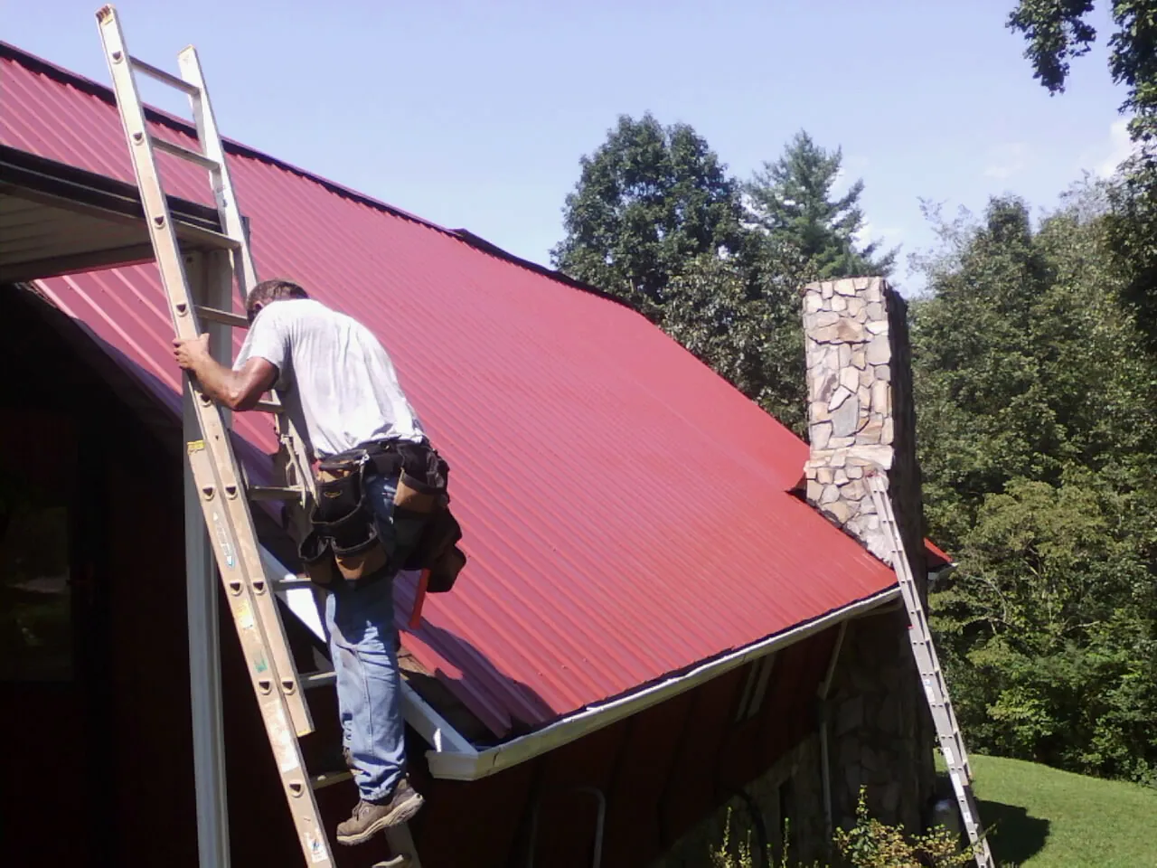 Roofer on a ladder beside a red metal roof and stone chimney — Soffit & Fascia Repair by Roofing Brothers in Haubstadt
